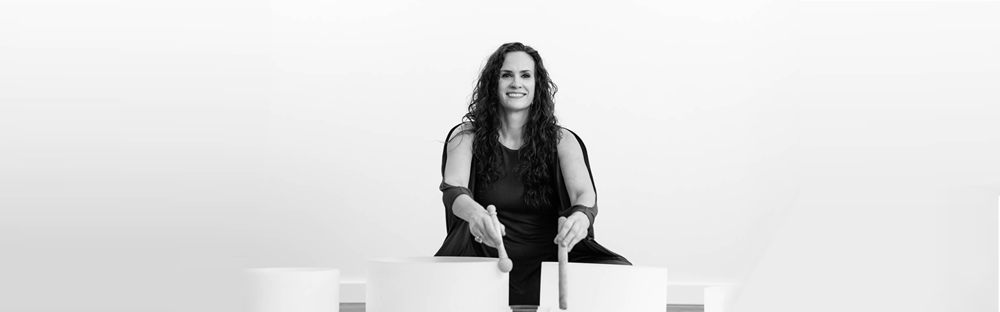 Image of a woman with dark wavy hair sitting behind several large crystal bowls as she plays them with mallet strikers.
