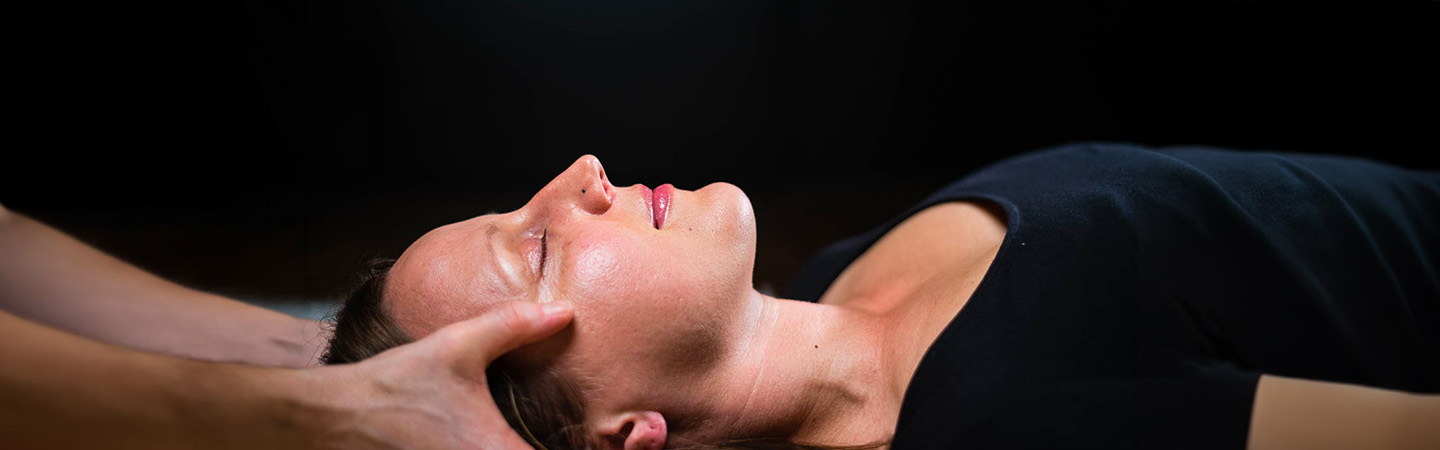 Image a woman laying with eyes closed while hands of a bodywork hold both sides of her head gently.